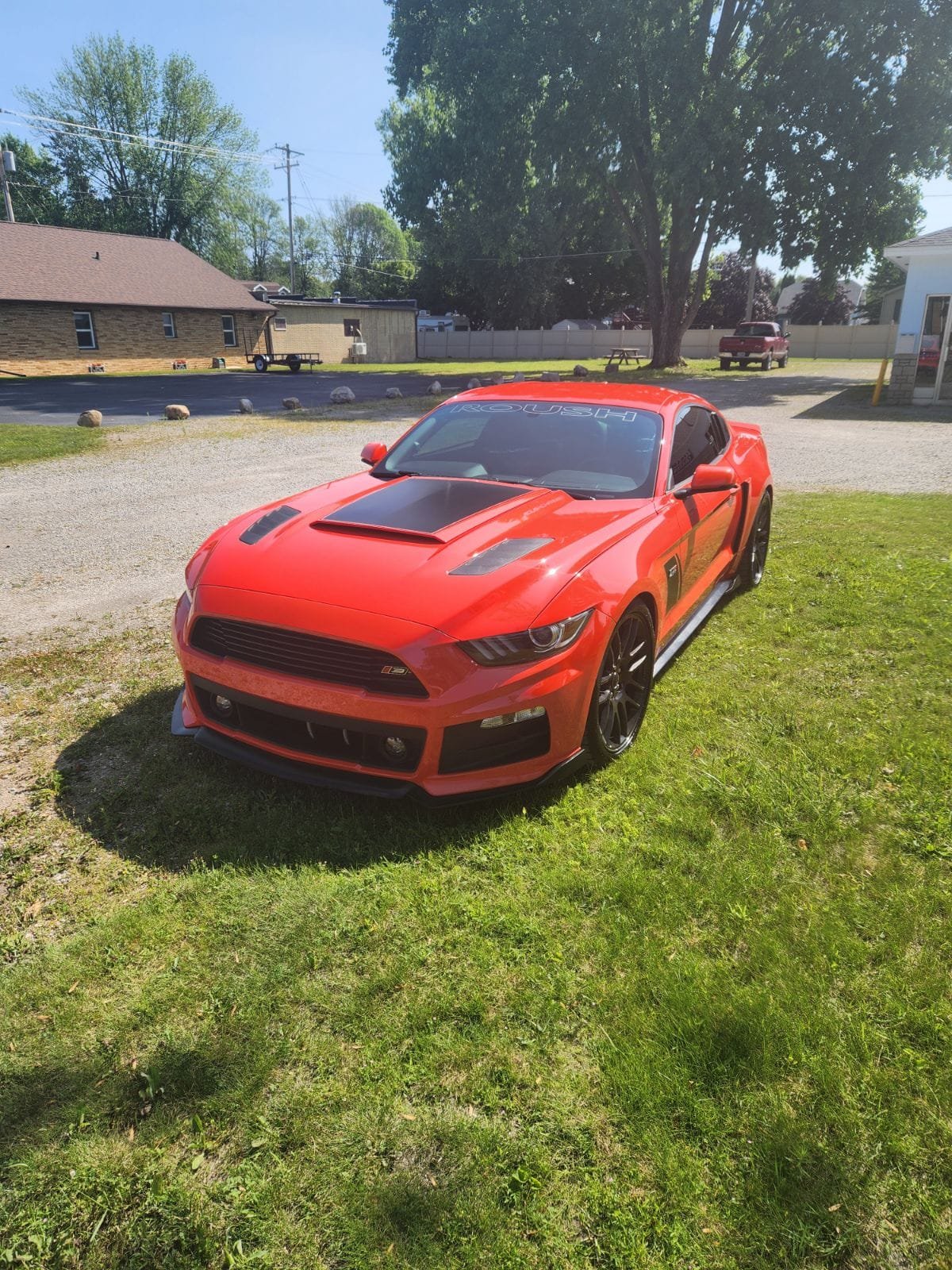 Red Mustang sports car parked on grass in front of rural buildings and large trees
