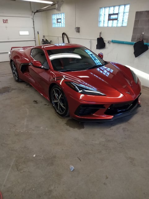 Red sports car parked in a concrete garage with white walls, fluorescent lighting, and an exit sign visible